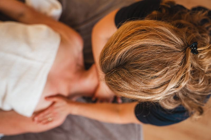 Therapeutin steht am Kopfende einer Massageliege und dehnt die fasziale Hals-und Nackenstrucktur eine auf dem Rücken liegenden Frau