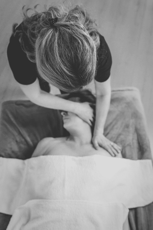 Therapeutin steht am Kopfende einer Liege und dehnt die fasziale Hals-und Nackenstruktur einer auf dem Rücken liegenden Frau