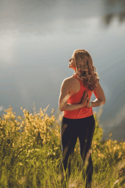 eine Frau von hinten mit geschlossenen Augen im sportlichen Outfit mit gefalteten Handflächen hinter dem Rücken, das Gesicht zur Seite der Sonne zugewandt