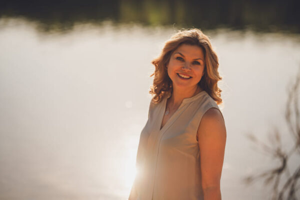 obere Hälfte einer lächelnden Frau, vor einem See als Hintergrung und einer leichten Sonnenspiegelung