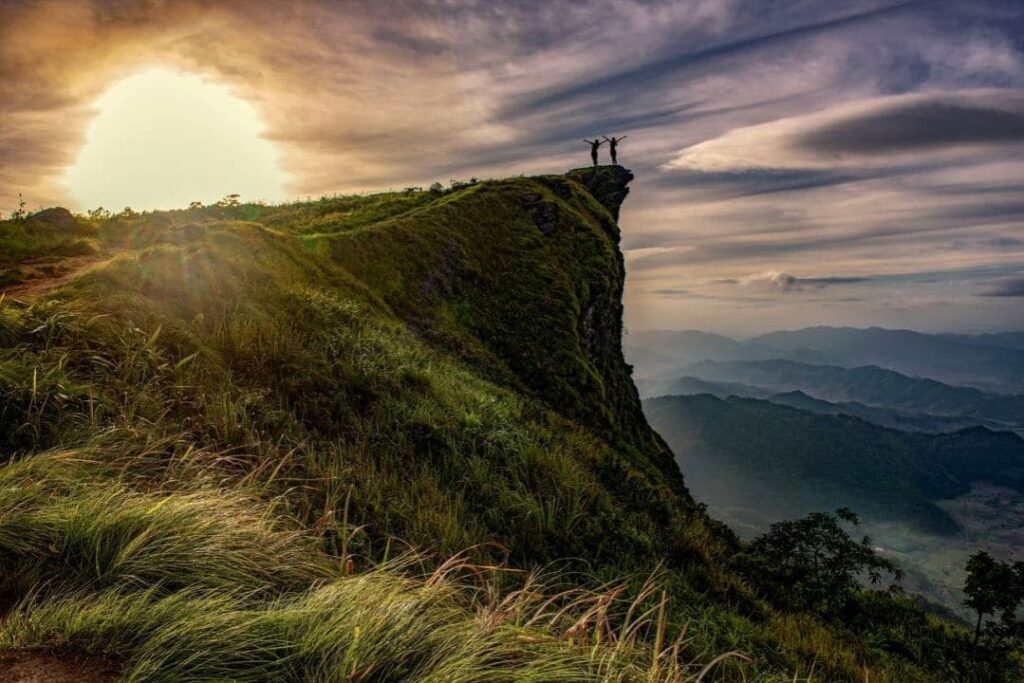 ein landschaftliches Panoramabild mit Sonnenaufgang und einem Berg auf dessen Spitze zwei menschliche Silhouetten ihre Hände in die Luft zur Seite heben